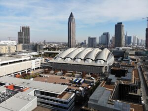 Aerial view of a city with modern skyscrapers and the distinctive building featuring curved white roofs, surrounded by urban streets and smaller buildings under a blue sky.