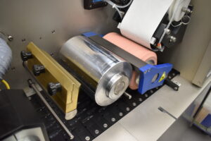 Close-up of an Ackley Hartnett printing press machine showing a large metal roller, a pink rubber roller, and a roll of white paper, with various metal mechanical parts and components visible.