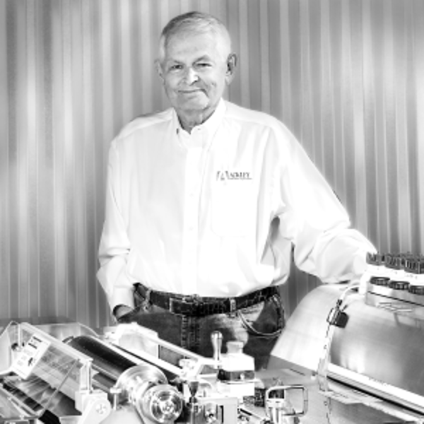 Michael Ackley Founder, in a white button-up shirt stands with one hand resting on industrial equipment, smiling in front of a corrugated metal wall.
