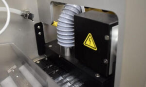 Close-up of industrial machinery with a flexible hose, black box with a yellow electrical hazard warning symbol, and metal and plastic components, possibly part of a manufacturing or processing machine.
