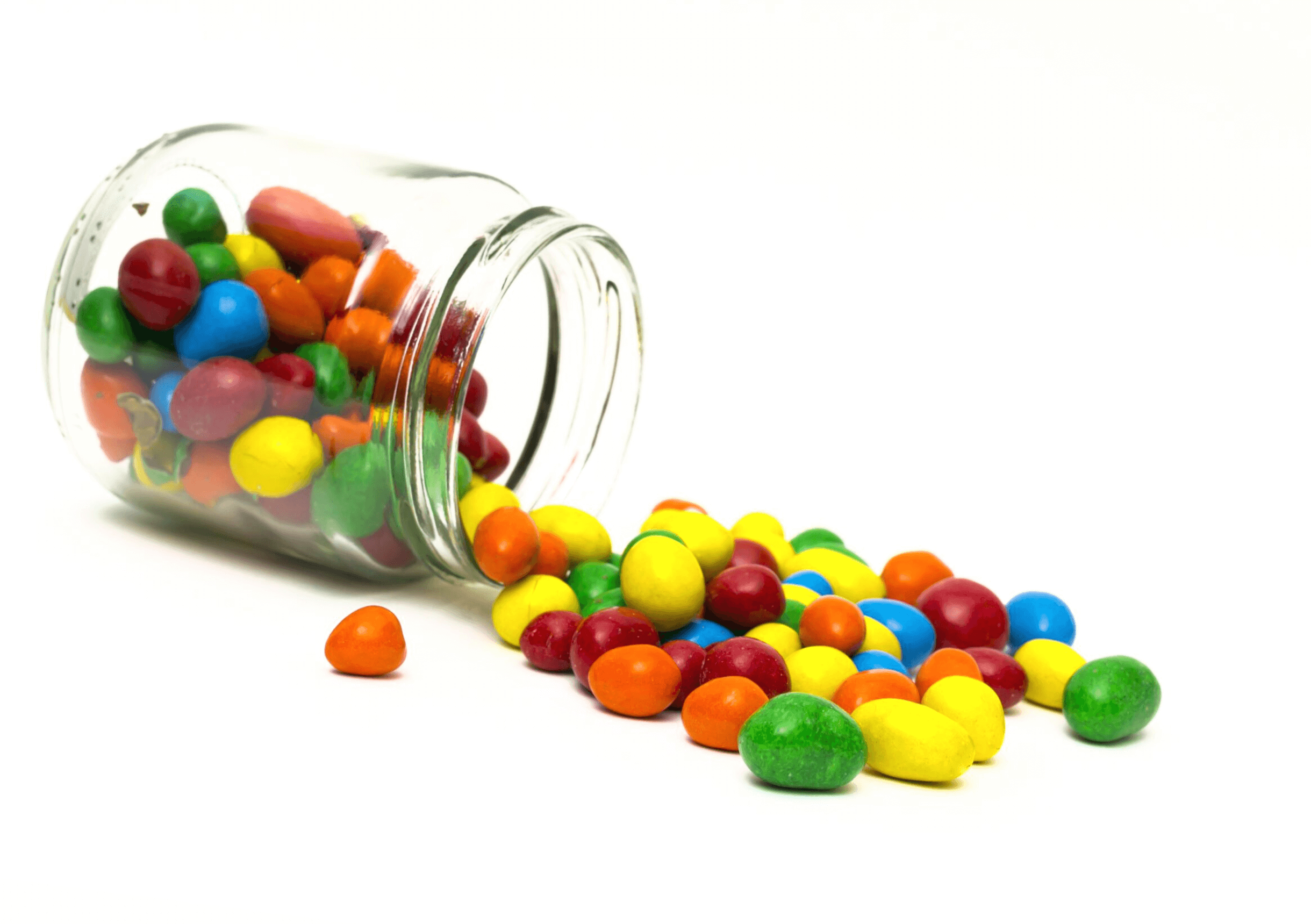 A glass jar tipped over on its side, spilling colorful candy-coated chocolates onto a white surface.