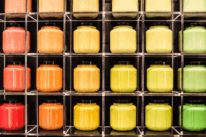 A grid of colorful jars organized in clear cubby shelves, featuring shades of red, orange, yellow, and green edible inks with black lids—each row displays different types of edible ink, neatly lined up in vibrant harmony.