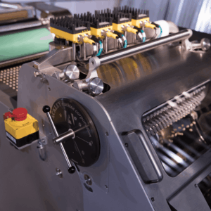 A close-up of an industrial printing press with a control panel, hand crank, emergency stop button, and rollers, showing mechanical and electronic components.