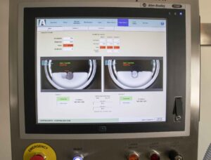 A touchscreen monitor displays a vision inspection system for pharmaceuticals with statistics, graphs, and live camera feeds of a production line. Below the screen are emergency, reset, and laser control buttons.
