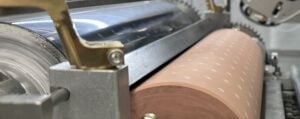 Close-up view of a a scraper blade with metal gears and rollers, including a copper-colored roller with a pattern of small white markings, likely used in an industrial or manufacturing process.