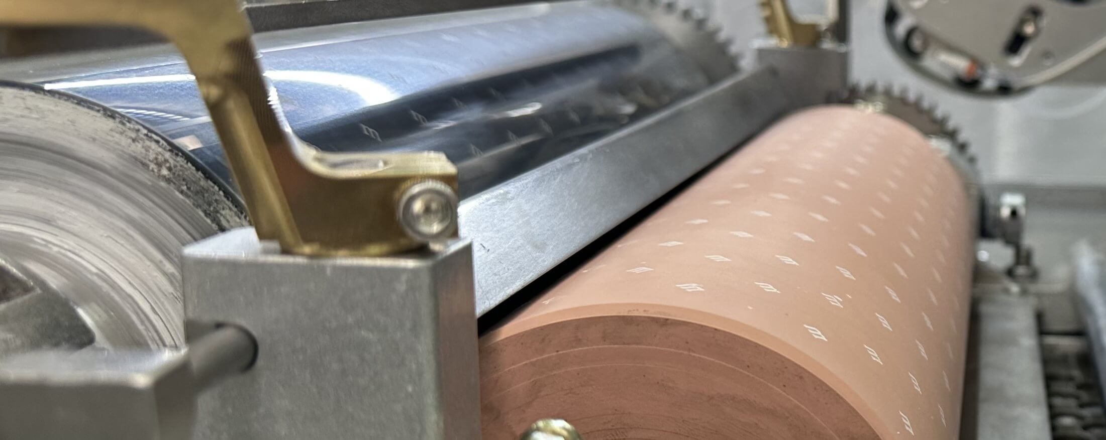 Close-up view of a a scraper blade with metal gears and rollers, including a copper-colored roller with a pattern of small white markings, likely used in an industrial or manufacturing process.