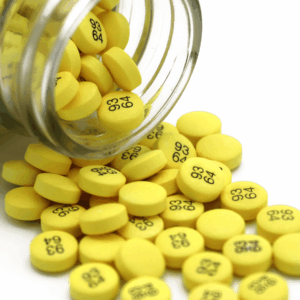 A glass bottle tipped over, spilling numerous round yellow pills with black "93 64" capsule printing with a white background.