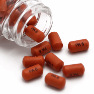 A clear bottle tipped over, spilling several small, cylindrical, dark orange tablets labeled "PA 6" with crisp capsule printing onto a white surface.