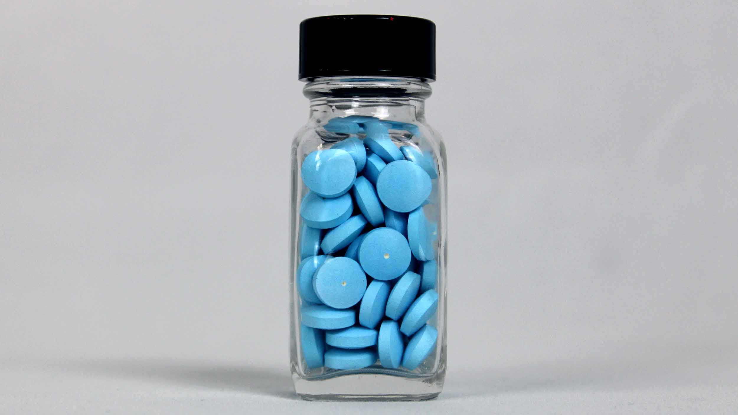 A clear glass bottle with a black cap, filled with round blue oral solid dosage tablets, is standing on a plain light gray surface with a neutral background.
