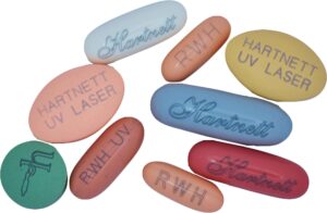 Assorted laser printing examples of colorful pills in various shapes and sizes, some labeled "HARTNETT UV LASER," "Hartnett," "RWH," and "UV."