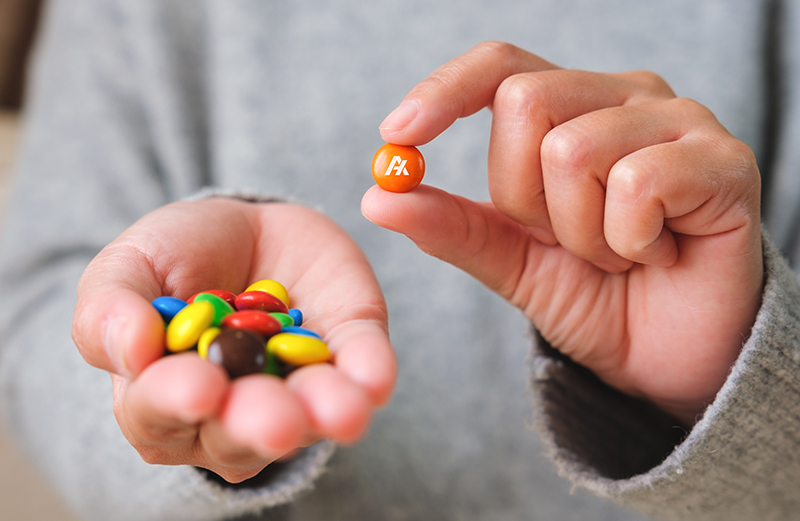 A person in a gray sweater holds colorful candy-coated chocolates, showing off the distinct candy marking as they pinch an orange one with the other hand.
